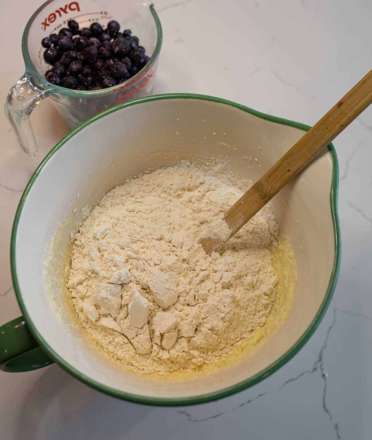 A mixing bowl with flour and a wooden spoon sits on a counter next to a measuring cup filled with blueberries, ready for baking delicious blueberry streusel muffins.