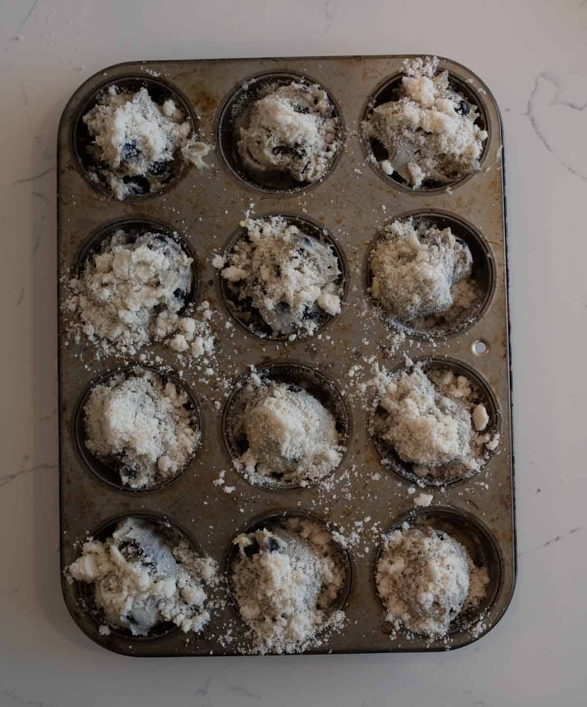 A muffin tin with twelve cups, each filled with blueberry streusel muffin batter topped with crumbly streusel, sitting on a light-colored marble countertop.