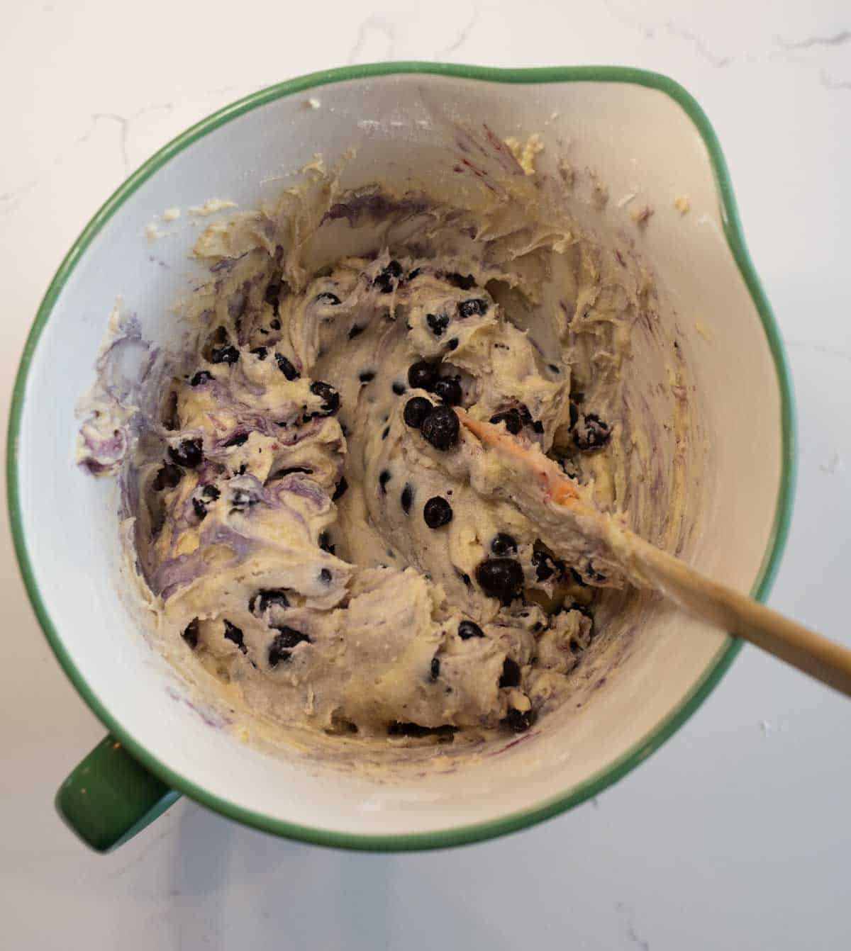 A green mixing bowl filled with blueberry streusel muffin batter, showing swirls of purple from the berries, with a wooden spoon resting inside on a white countertop.