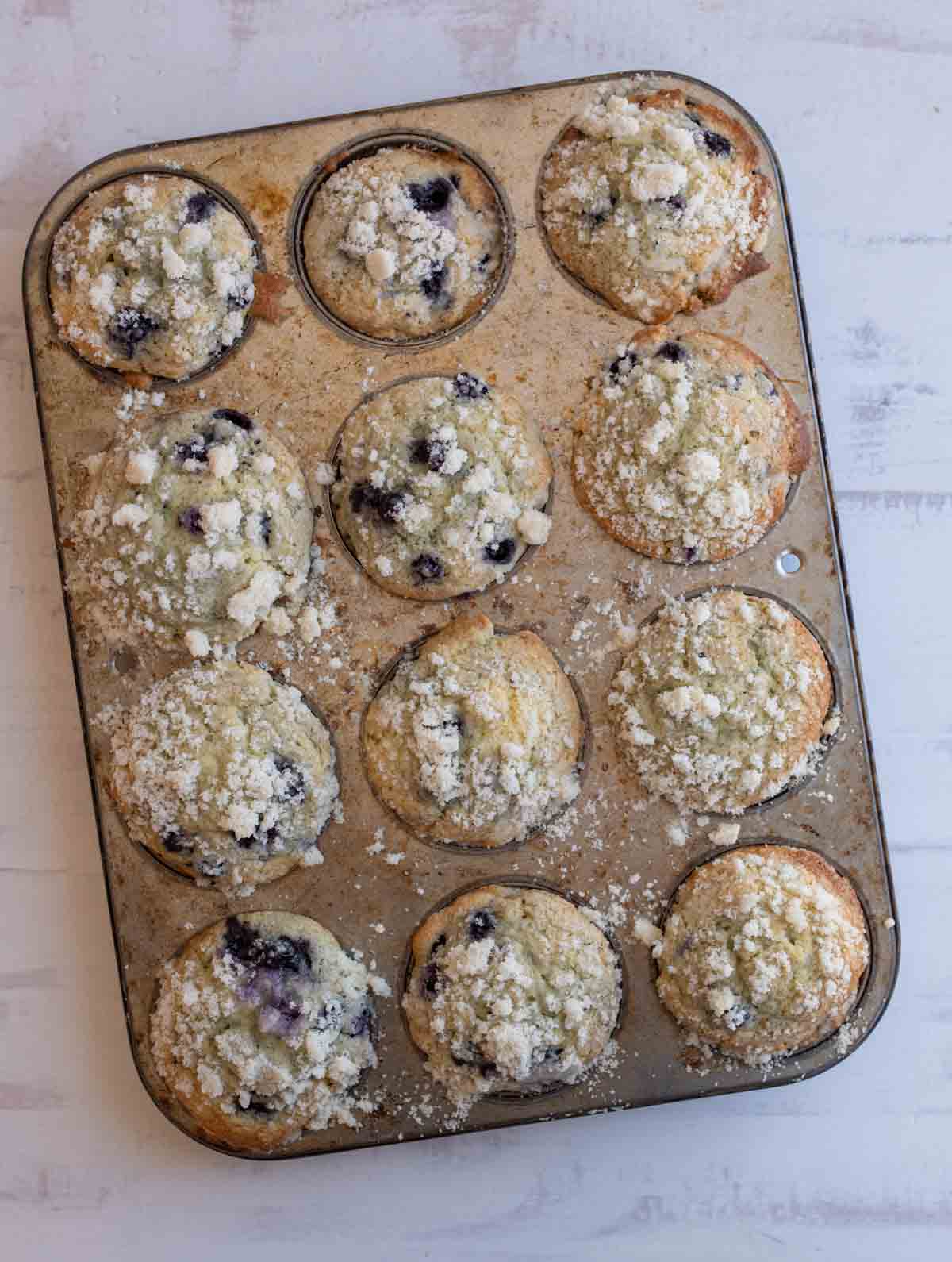 A muffin tin filled with twelve blueberry streusel muffins, their crumbly tops golden and uneven, sits on a light-colored surface—some muffins slightly overfilled for a rustic, homemade look.