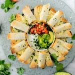 A round platter of baked chicken taquitos arranged in a circle around a bowl of sour cream, pico de gallo, guacamole, and avocado slices, garnished with shredded cheese and fresh cilantro.