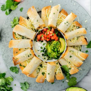 A round platter of baked chicken taquitos arranged in a circle around a bowl of sour cream, pico de gallo, guacamole, and avocado slices, garnished with shredded cheese and fresh cilantro.