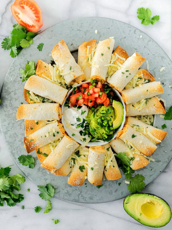 A round platter of baked chicken taquitos arranged in a circle around a bowl of sour cream, pico de gallo, guacamole, and avocado slices, garnished with shredded cheese and fresh cilantro.