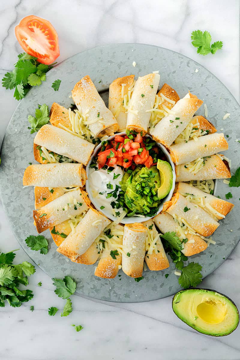A round platter of baked chicken taquitos arranged in a circle around a bowl of sour cream, pico de gallo, guacamole, and avocado slices, garnished with shredded cheese and fresh cilantro.