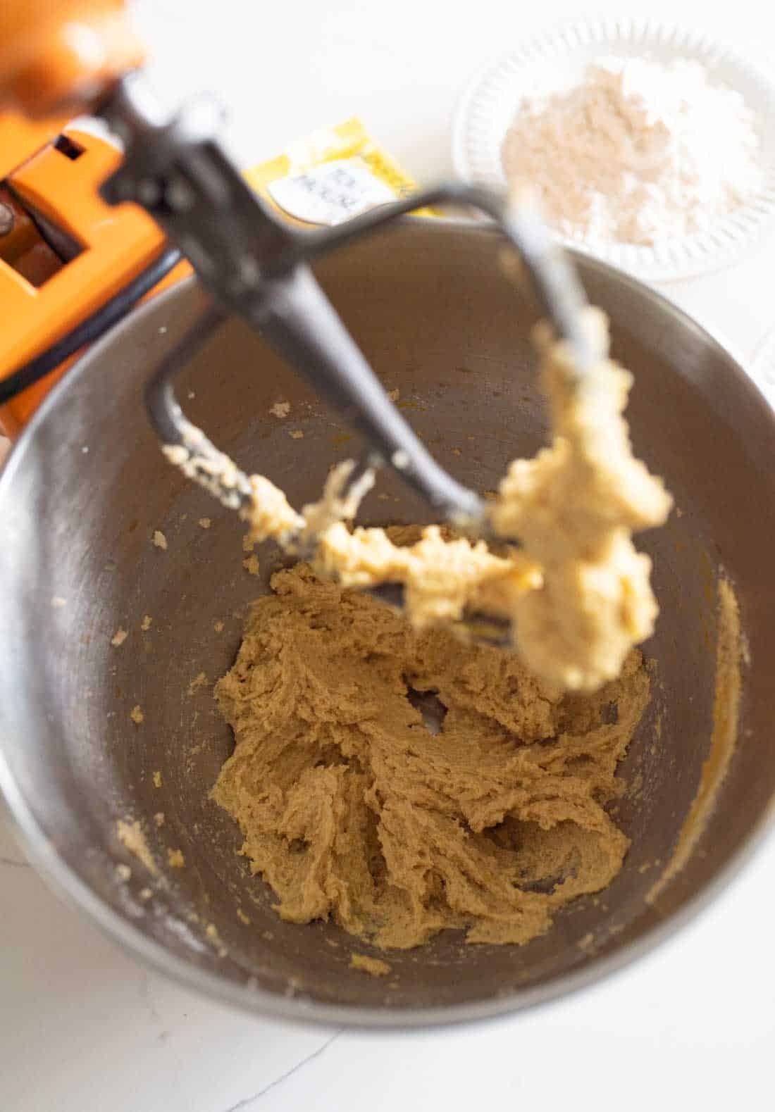 A mixing bowl with cookie dough being creamed by an electric mixer. In the background, flour on a plate and a packet of baking powder are visible on the counter.