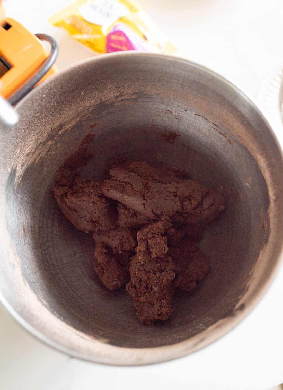 A metal mixing bowl containing dark brown chocolate cookie dough, with a stand mixer attachment partially visible and a bag of sugar in the background.