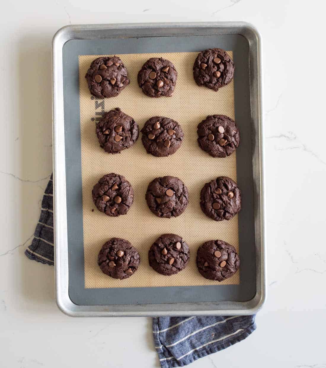 A baking sheet with twelve chocolate cookies topped with chocolate chips, arranged in rows of three on a silicone mat, sits on a white surface with a striped kitchen towel partially underneath.