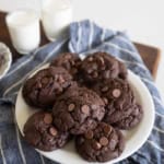 A white plate of double chocolate chip cookies sits on a blue striped cloth, with two glasses of milk in the background. The cookies are dark and studded with chocolate chips.