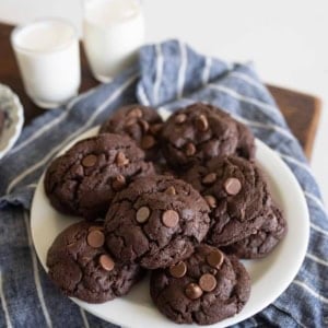 A white plate of double chocolate chip cookies sits on a blue striped cloth, with two glasses of milk in the background. The cookies are dark and studded with chocolate chips.