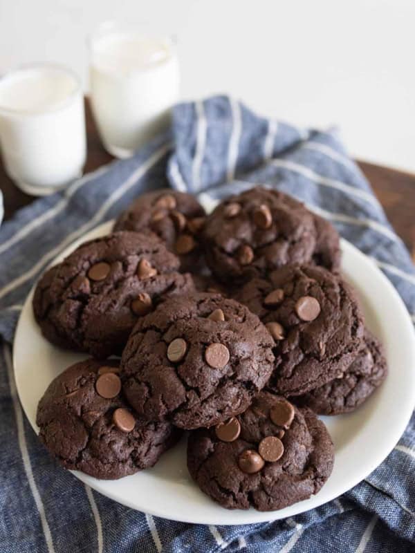 A white plate of double chocolate chip cookies sits on a blue striped cloth, with two glasses of milk in the background. The cookies are dark and studded with chocolate chips.