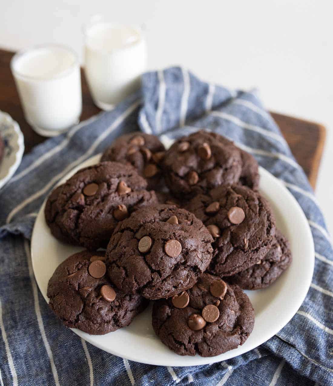 A white plate of double chocolate chip cookies sits on a blue striped cloth, with two glasses of milk in the background. The cookies are dark and studded with chocolate chips.