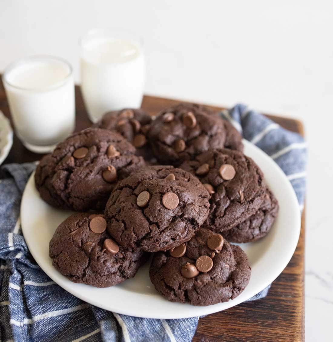 A white plate filled with chocolate chocolate-chip cookies sits on a blue and white cloth, with two glasses of milk in the background on a wooden surface.