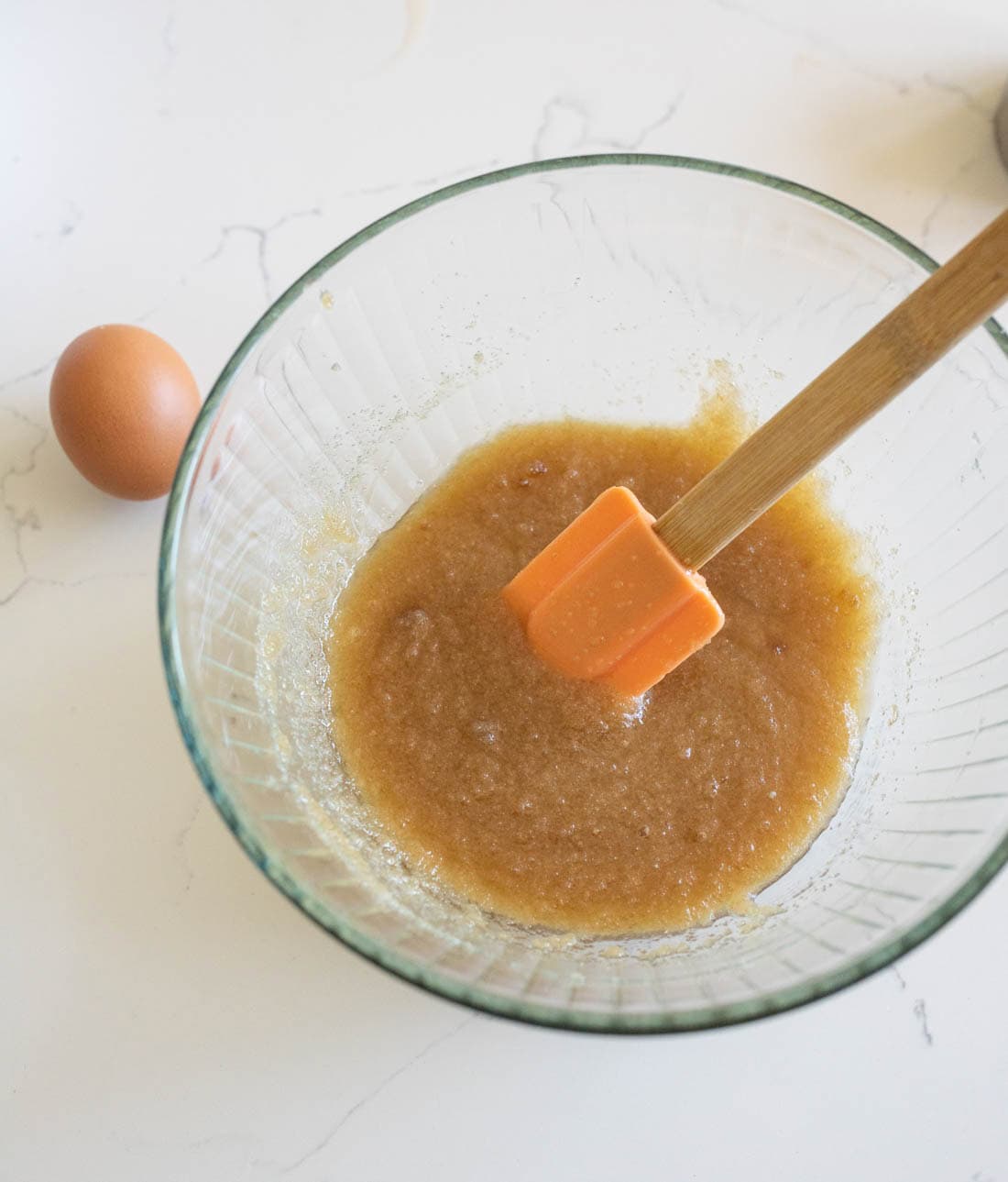 A glass bowl with a light brown mixture being stirred by an orange spatula sits on a white countertop next to a whole brown egg.