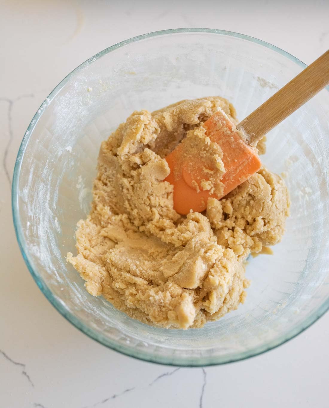 A glass bowl filled with cookie dough being mixed with an orange spatula, placed on a white countertop.