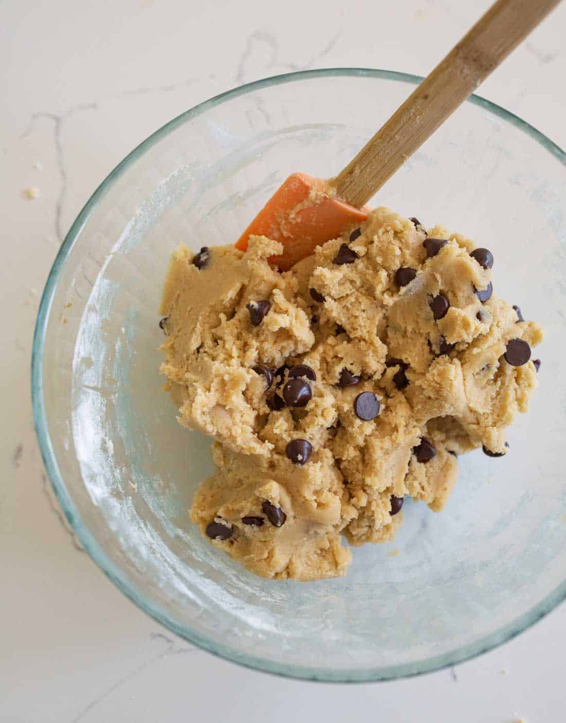 A glass bowl filled with chocolate chip cookie dough sits on a white countertop. A spatula with an orange head and wooden handle is resting inside the bowl.