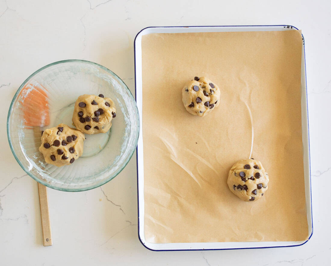 A glass bowl with chocolate chip cookie dough sits next to a baking tray lined with parchment paper. Three dough balls studded with chocolate chips are on the tray, ready for baking. A spatula rests in the bowl.