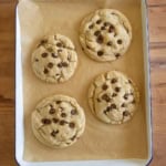 Four large chocolate chip cookies on brown parchment paper in a rectangular baking tray, placed on a wooden surface.