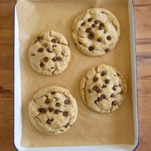 Four large chocolate chip cookies on brown parchment paper in a rectangular baking tray, placed on a wooden surface.