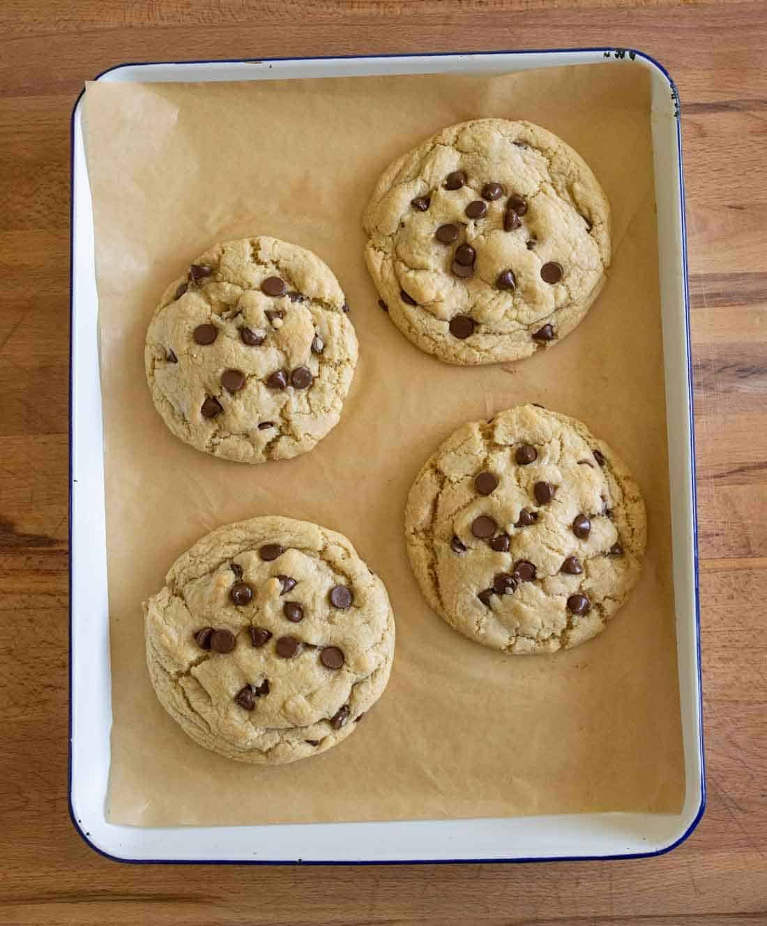 Four large chocolate chip cookies on brown parchment paper in a rectangular baking tray, placed on a wooden surface.