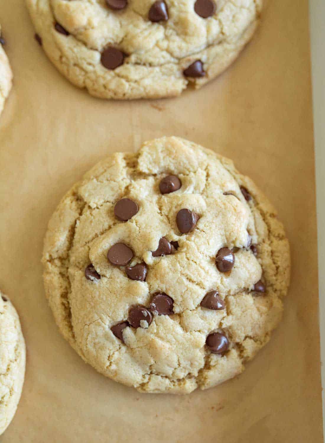 A close-up of a freshly baked chocolate chip cookie with a golden-brown, slightly cracked surface and several melted chocolate chips, resting on parchment paper.