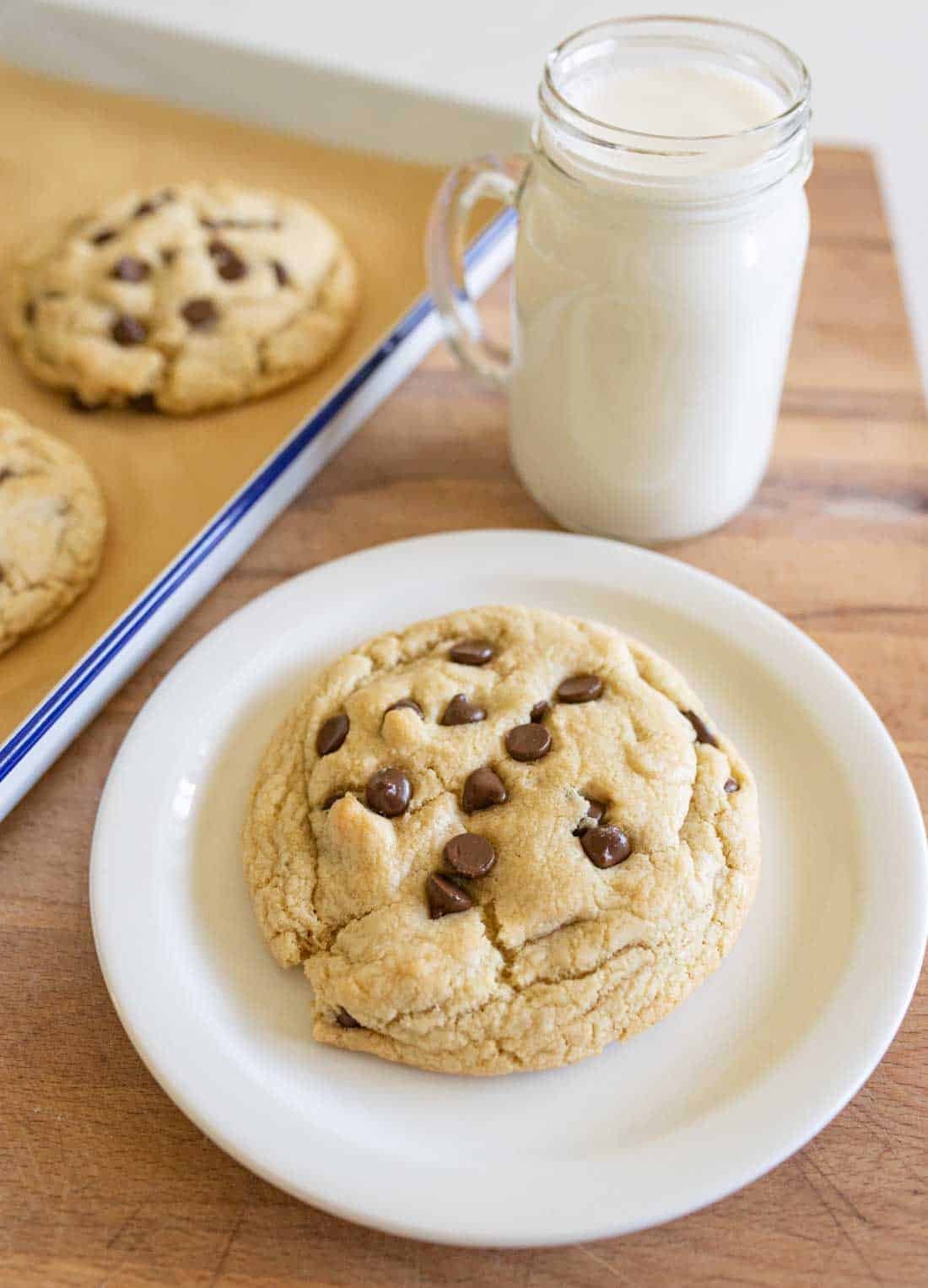 A large chocolate chip cookie on a white plate sits next to a glass jar of milk. Additional cookies on a baking tray are visible in the background made from a giant chocolate chip cookie recipe.