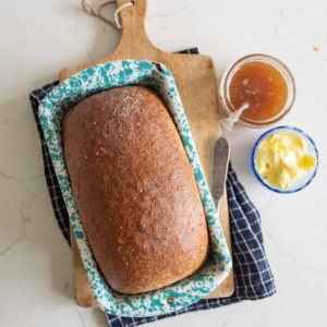 A loaf of King Arthur Flour whole wheat bread in a blue speckled pan sits on a wooden cutting board with a knife, next to a jar of jam and a small bowl of butter, all on a white surface with a dark checkered cloth.
