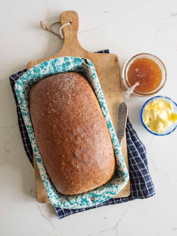 A loaf of King Arthur Flour whole wheat bread in a blue speckled pan sits on a wooden cutting board with a knife, next to a jar of jam and a small bowl of butter, all on a white surface with a dark checkered cloth.