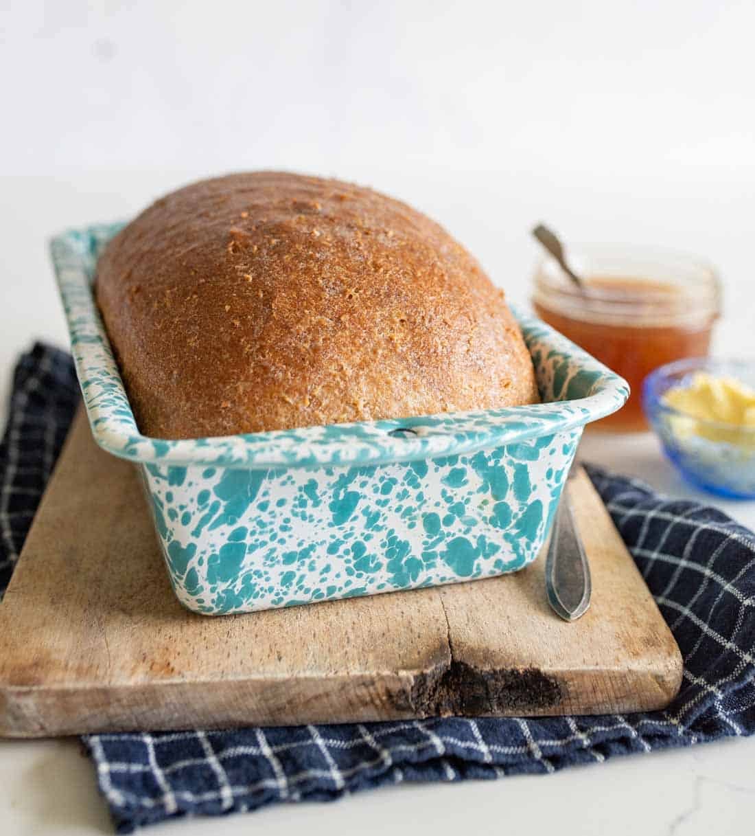 A loaf of bread in a blue and white speckled baking pan sits on a wooden cutting board with a blue checkered cloth, next to a jar of honey, a butter dish, and a spoon.