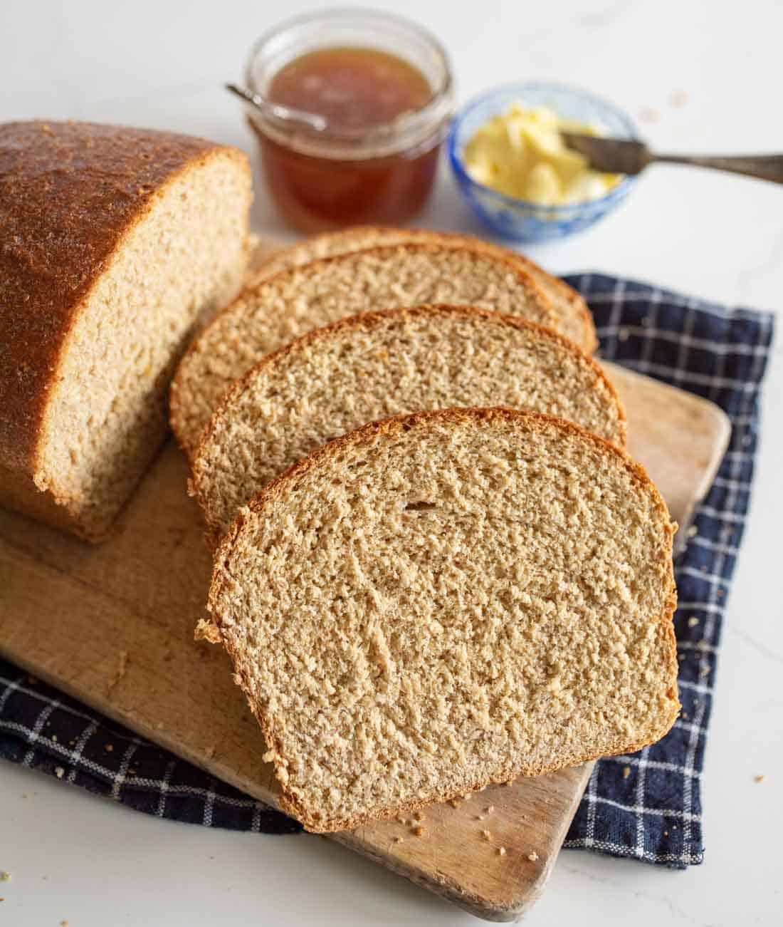 Three slices of whole wheat bread on a wooden cutting board with a loaf, a jar of honey, a dish of butter, and a butter knife on a blue plaid cloth.