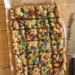 A batch of monster cookie bars topped with colorful candy-coated chocolates, cut into squares on parchment paper, with a large knife next to them on a wooden surface.