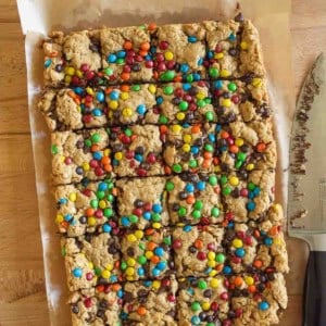 A batch of monster cookie bars topped with colorful candy-coated chocolates, cut into squares on parchment paper, with a large knife next to them on a wooden surface.