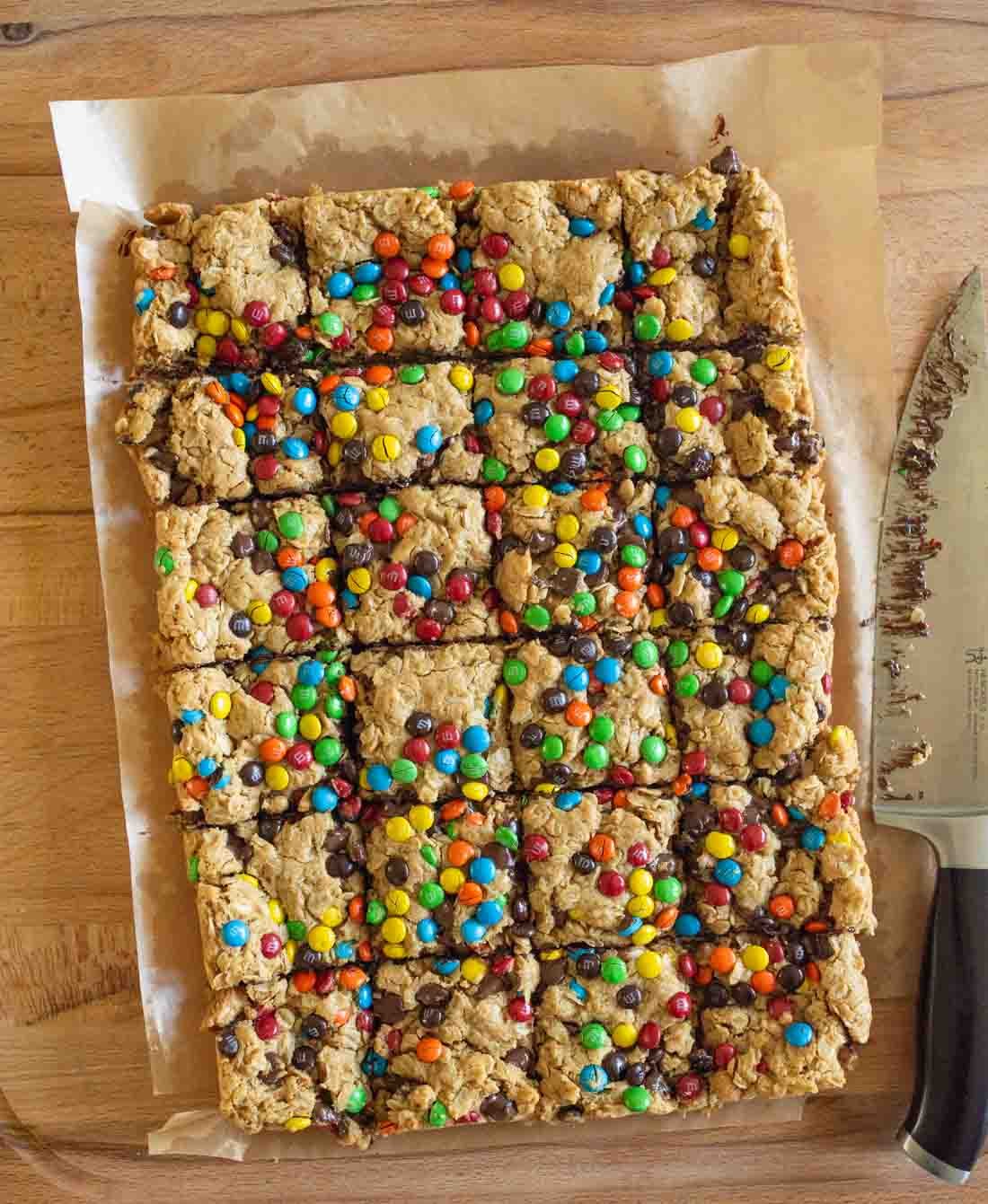 A batch of monster cookie bars topped with colorful candy-coated chocolates, cut into squares on parchment paper, with a large knife next to them on a wooden surface.
