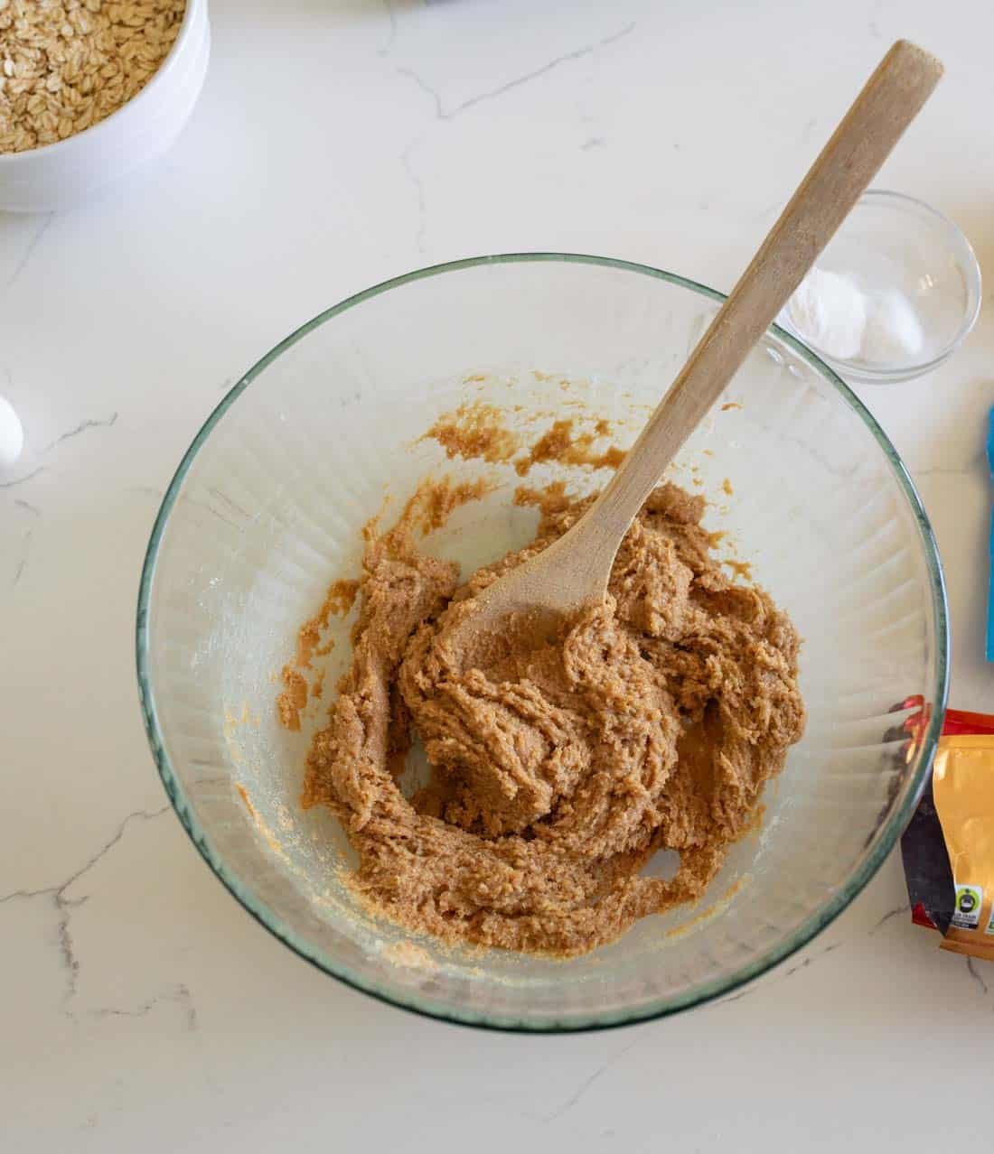 A glass mixing bowl with brown cookie dough and a wooden spoon on a white countertop, surrounded by a bowl of oats, a small bowl of white powder, and a partially visible bag of chocolate chips.