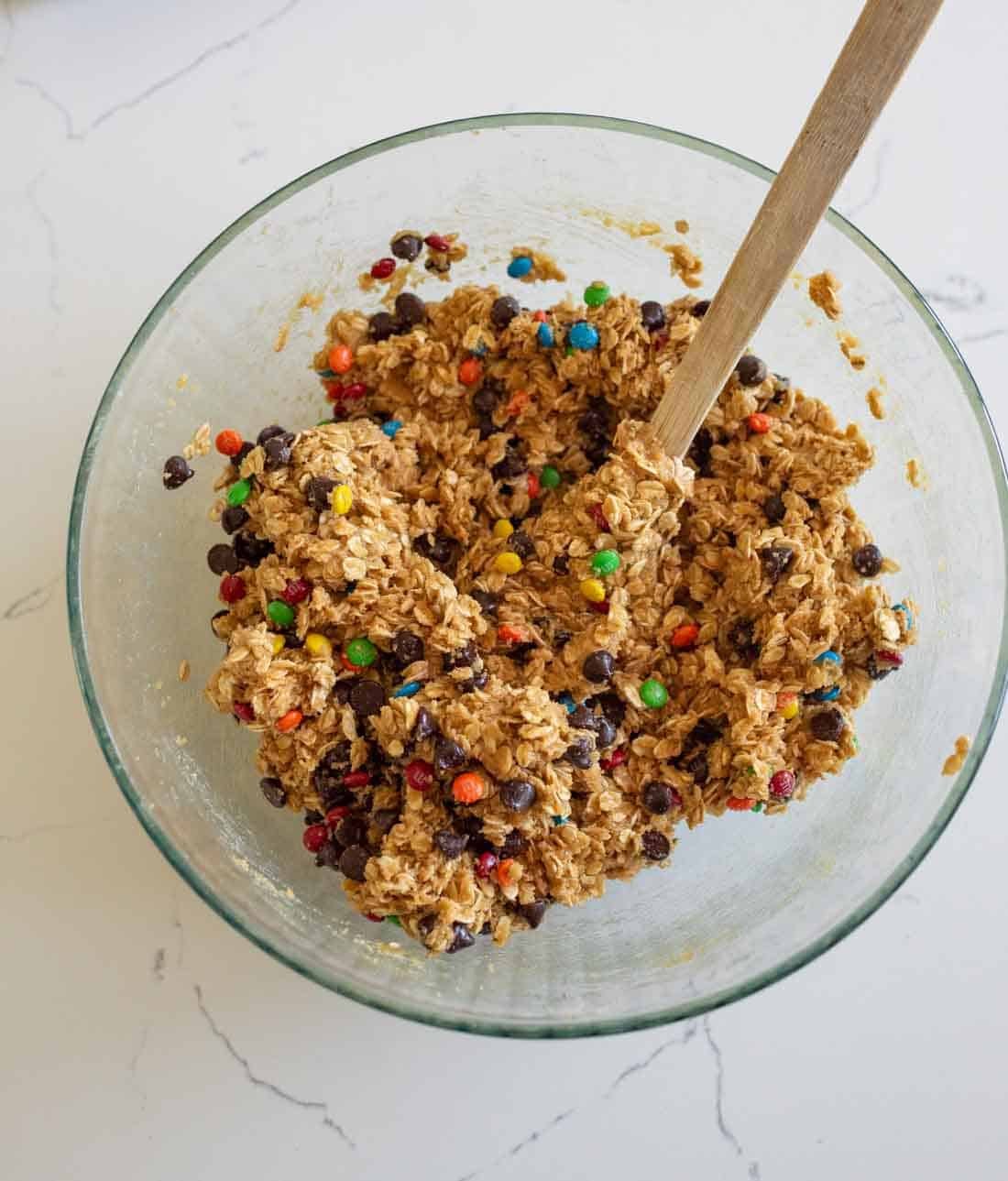 A glass bowl filled with cookie dough mixed with oats, chocolate chips, and colorful candy-coated chocolates, with a wooden spoon resting inside.