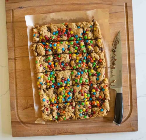 A batch of cookie bars topped with colorful candy-coated chocolates, cut into squares on parchment paper, sits on a wooden cutting board next to a knife.
