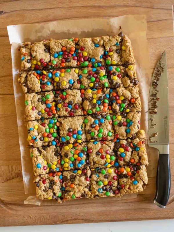 A batch of cookie bars topped with colorful candy-coated chocolates, cut into squares on parchment paper, sits on a wooden cutting board next to a knife.