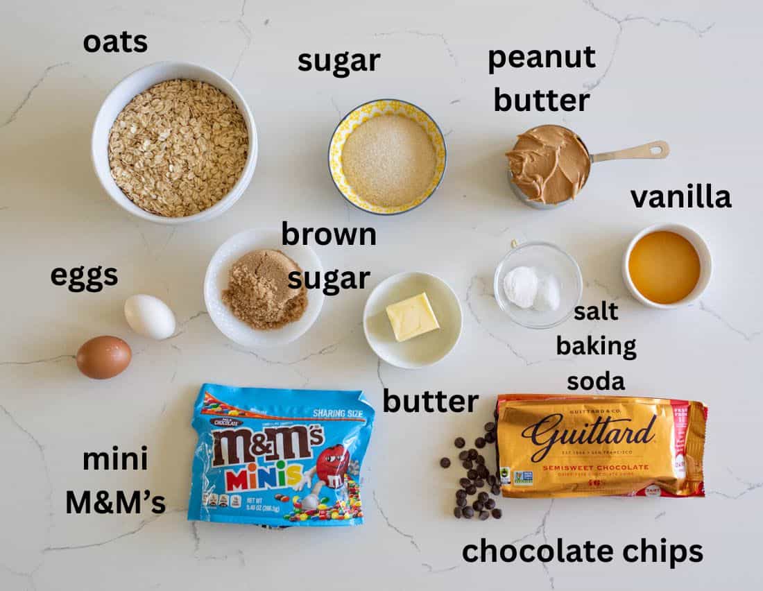 Ingredients for baking are arranged on a countertop and labeled, including oats, sugar, brown sugar, eggs, mini M&M’s, peanut butter, vanilla, salt, baking soda, butter, and chocolate chips.