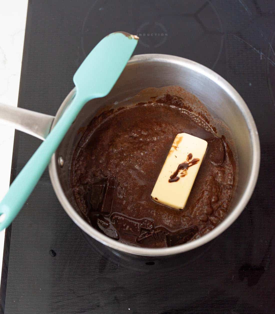 A saucepan on a stovetop contains partially melted butter and a chocolate mixture, with a blue spatula resting on the rim.