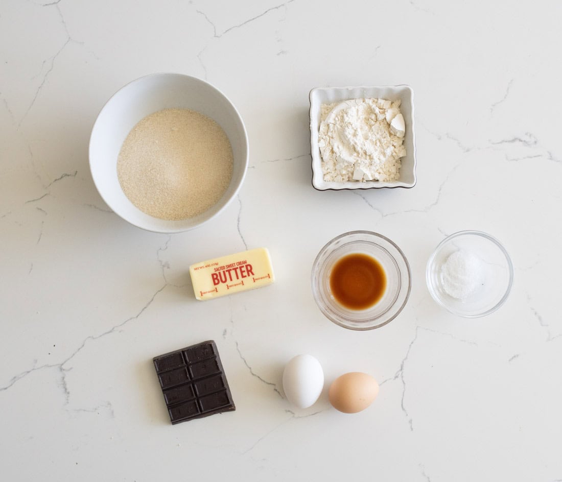A flat lay of baking ingredients on a white marble surface: a bowl of sugar, a dish of flour, a stick of butter, a small bowl of vanilla extract, a small bowl of salt, a bar of chocolate, a white egg, and a brown egg.