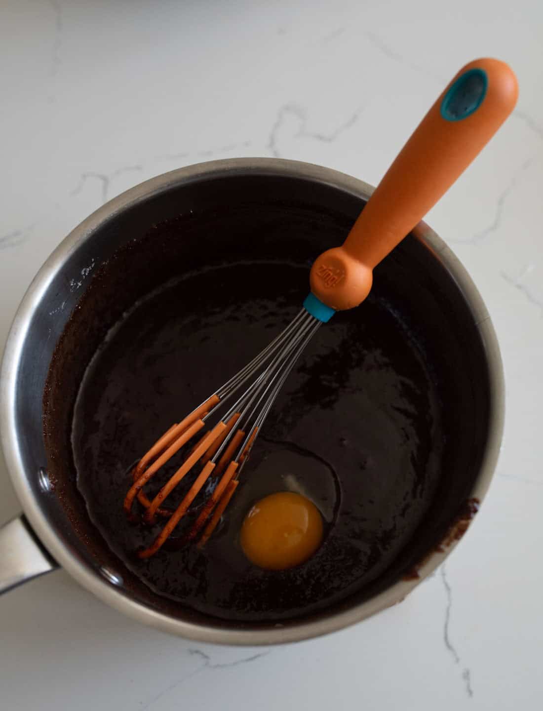 A saucepan with chocolate batter and a raw egg in the center, being mixed with an orange and blue whisk, sits on a white marble surface.