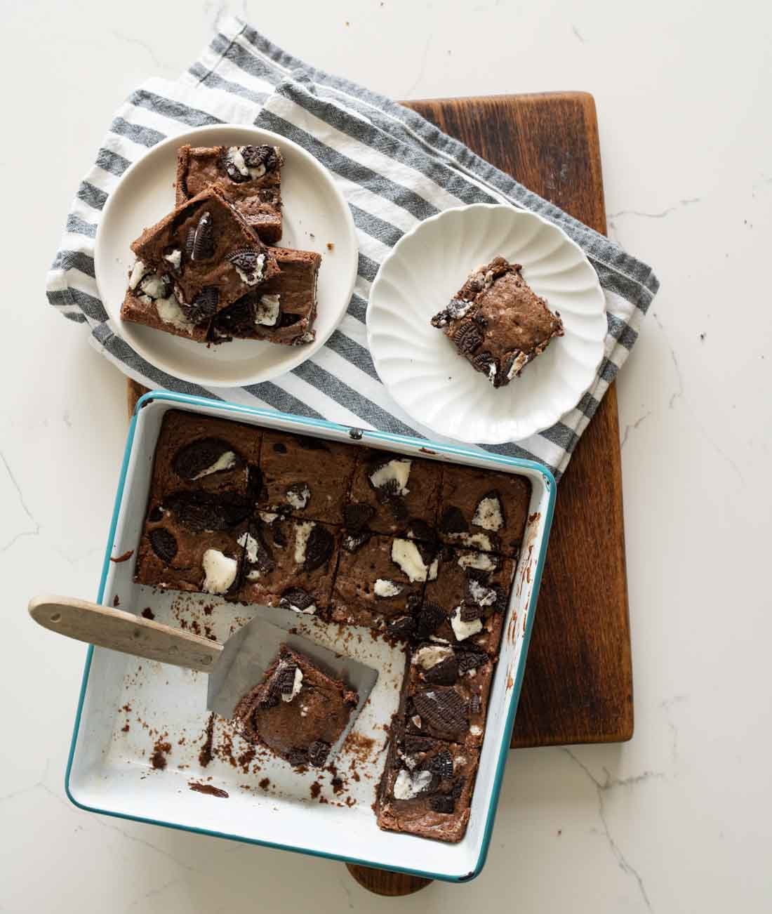A white baking dish with brownies, some cut and some removed, sits on a wooden board. A plate and a bowl nearby hold more brownies. A striped cloth is underneath the plate and bowl.