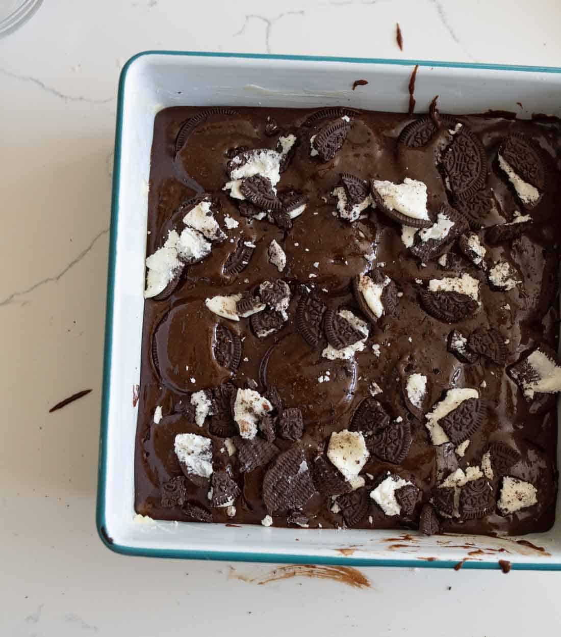 A square baking dish filled with chocolate brownie batter topped with crumbled Oreo cookies, sitting on a white countertop.