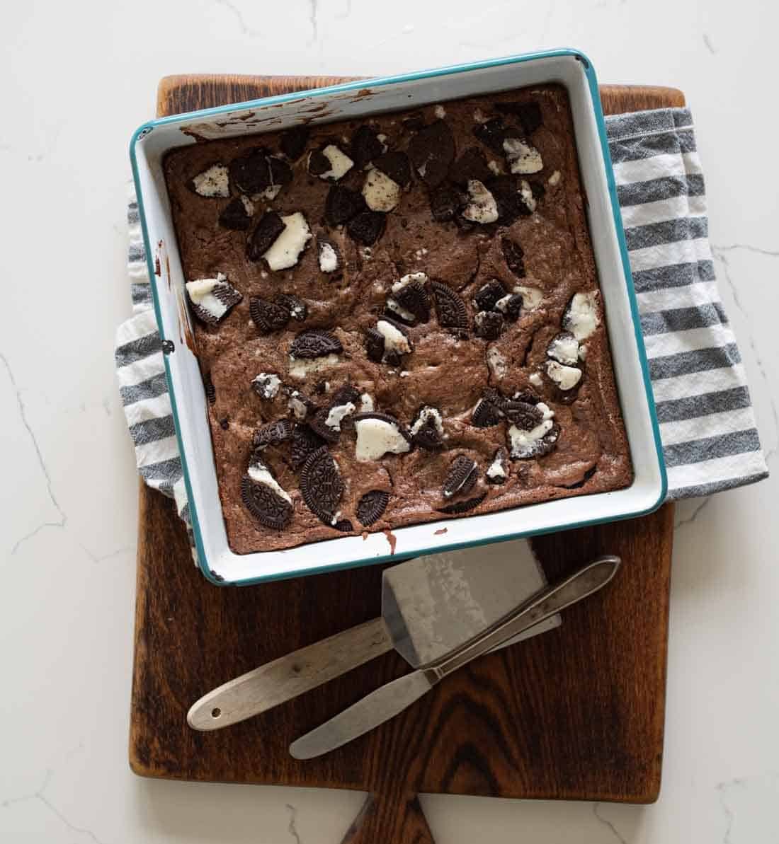 A pan of chocolate brownies topped with pieces of cookies sits on a wooden cutting board, next to a striped kitchen towel and metal serving utensils.