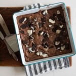A square baking pan filled with chocolate oreo brownies topped with broken Oreo cookies, resting on a striped kitchen towel and a wooden cutting board; a knife and a metal spatula are beside the pan.