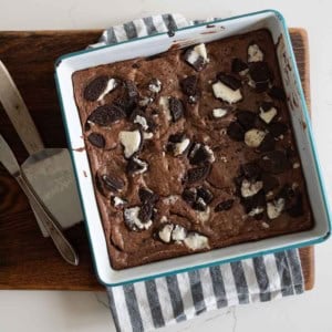 A square baking pan filled with chocolate oreo brownies topped with broken Oreo cookies, resting on a striped kitchen towel and a wooden cutting board; a knife and a metal spatula are beside the pan.