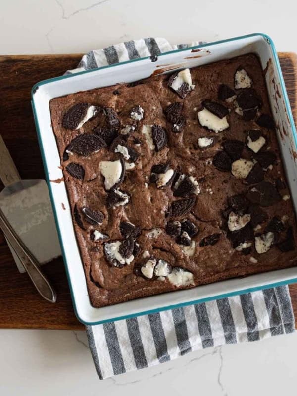 A square baking pan filled with chocolate oreo brownies topped with broken Oreo cookies, resting on a striped kitchen towel and a wooden cutting board; a knife and a metal spatula are beside the pan.