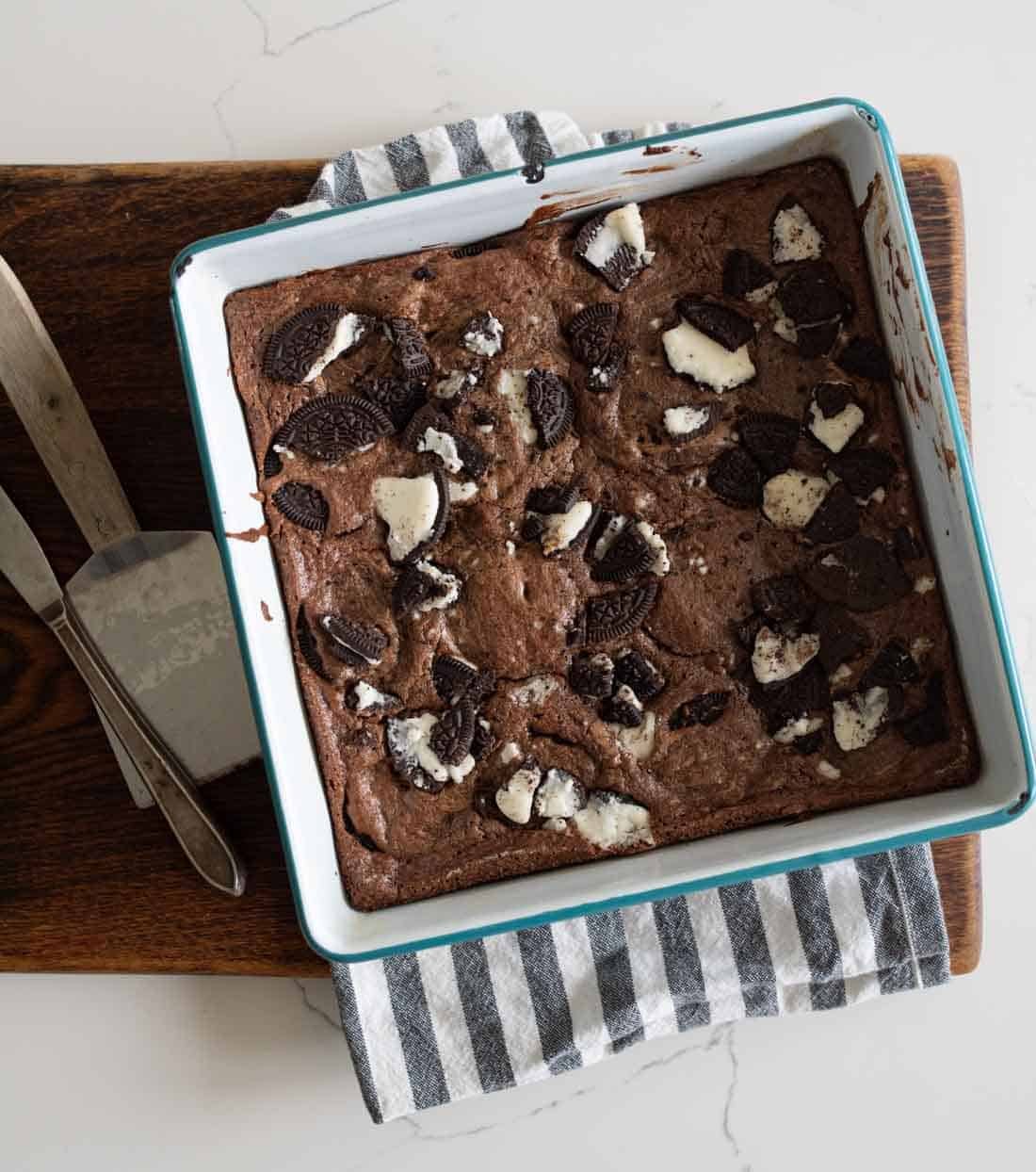 A square baking pan filled with chocolate oreo brownies topped with broken Oreo cookies, resting on a striped kitchen towel and a wooden cutting board; a knife and a metal spatula are beside the pan.