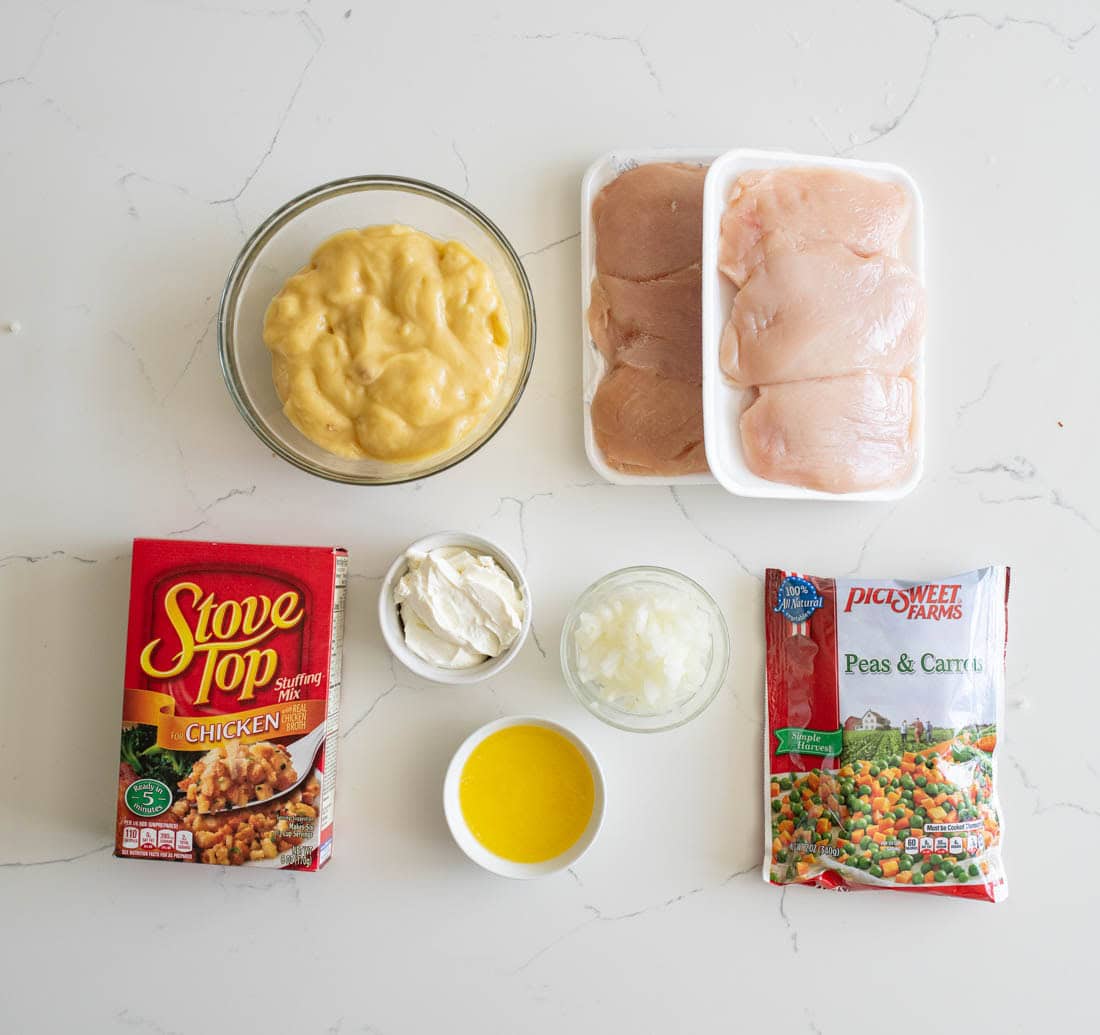 Ingredients on a white surface: raw chicken breasts in trays, a bowl of cream soup, boxed stuffing mix, a bag of frozen peas and carrots, a bowl of sour cream, a bowl of melted butter, and a bowl of chopped onions.