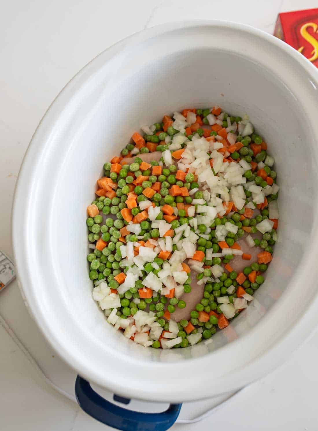 A slow cooker containing a mixture of chopped onions, carrots, and green peas. The vegetables are uncooked and evenly spread at the bottom of the slow cooker.