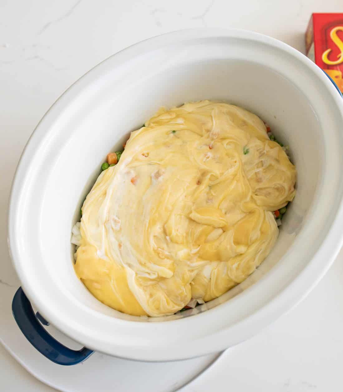 A white slow cooker filled with vegetables and topped with a thick layer of creamy yellow sauce, sitting on a white countertop. A partial box with the word Soup is visible in the corner.